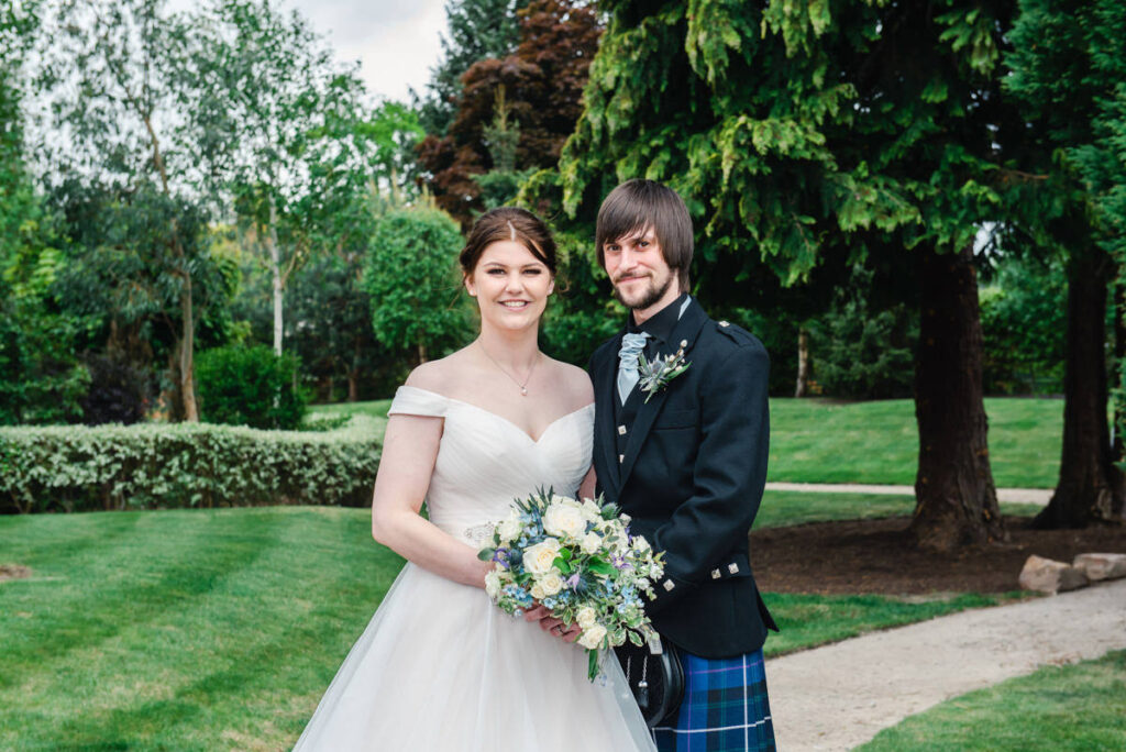 A bride in white and groom in blue kilt stand together holding a bouquet in the gardens of the Kingsmills hotel, Inverness