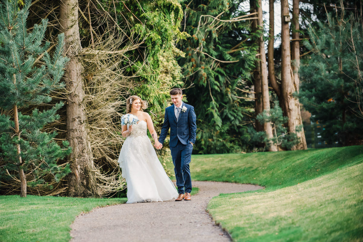 A bride and groom hold hands while walking along a woodland path in the gardens of the Kingsmills Hotel, Inverness