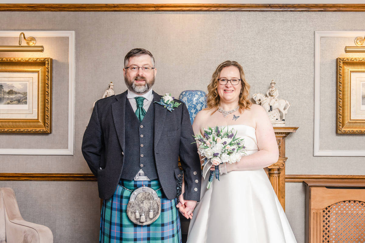 Bride and groom both wearing glasses stand in front of a fire place inside the Kingsmills hotel , Inverness