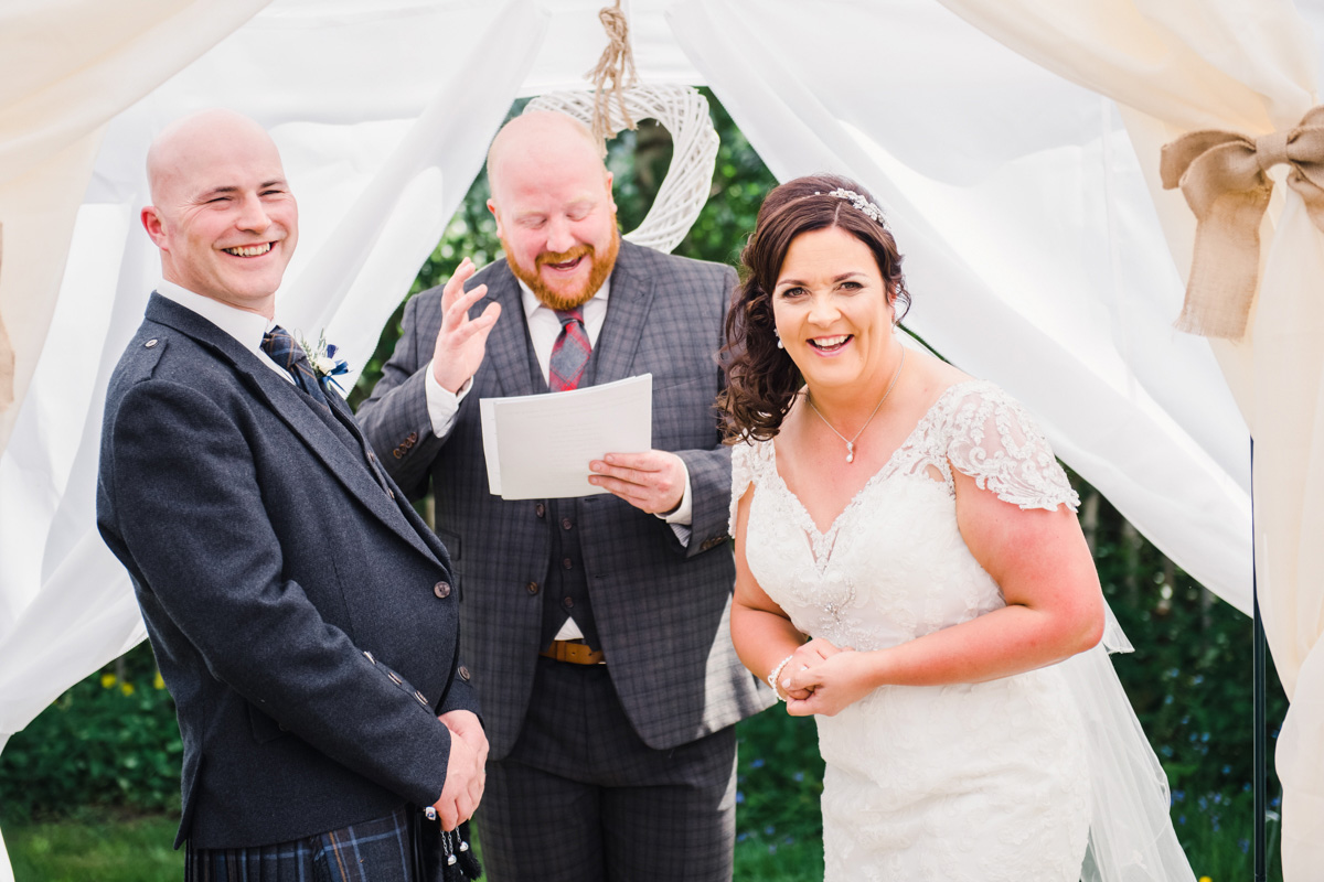 Bride and groom laugh as a red bearded celebrant in cheque suit makes a joke in Loch Ness country house hotel near Inverness
