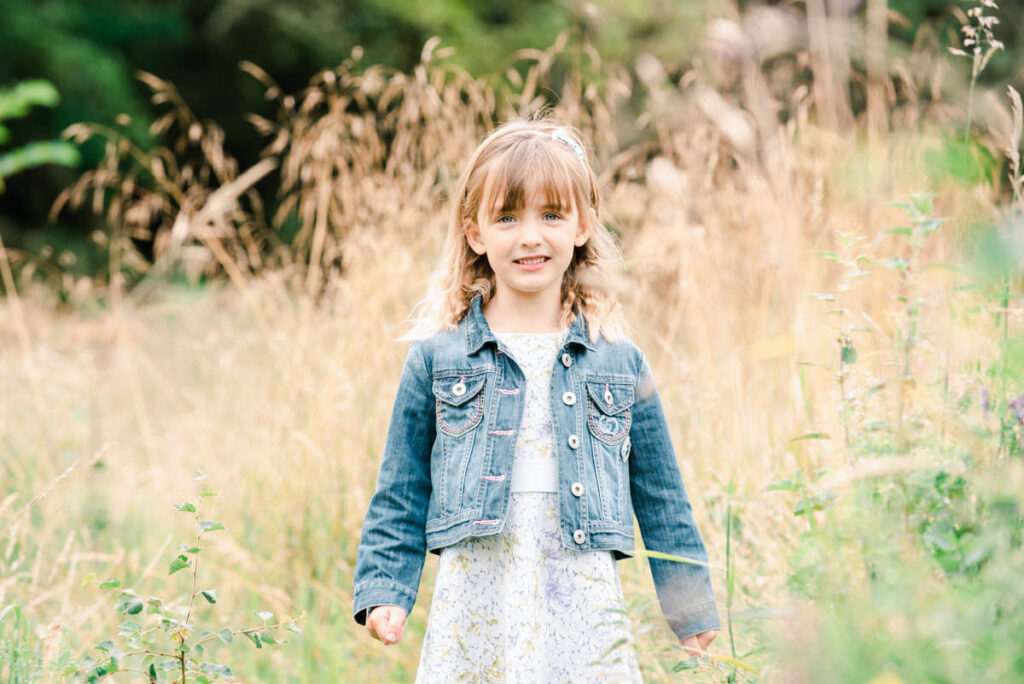 A young girl in a denim jacket and dress stands among tall grass during a Family Photo Shoot at Nairnside