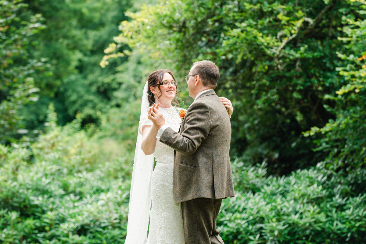 Bride in a veil and glasses dances with groom in beige suit outdoors surrounded by trees Pitcalzean house