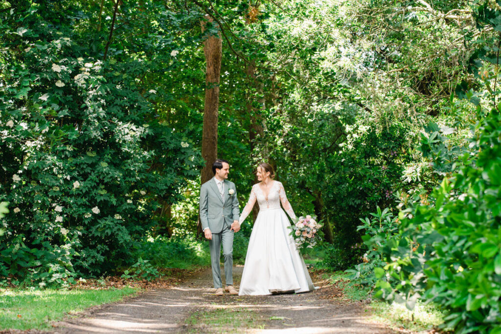 Bride holding a large bouquet walks hand in hand with the groom in a grey suit along a woodland track at Pitcalzean house