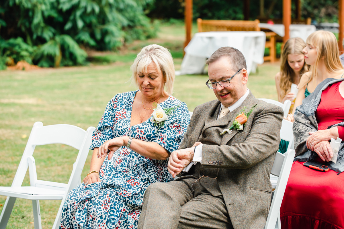 Man in a beige suite sits outside next to a woman in a blue floral dress checking the time at an outdoor wedding near Tain