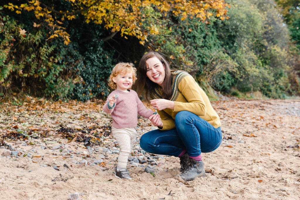 A red headed toddler stands next to her kneeling mother during a Family Photo Shoot on Rosemarkie beach