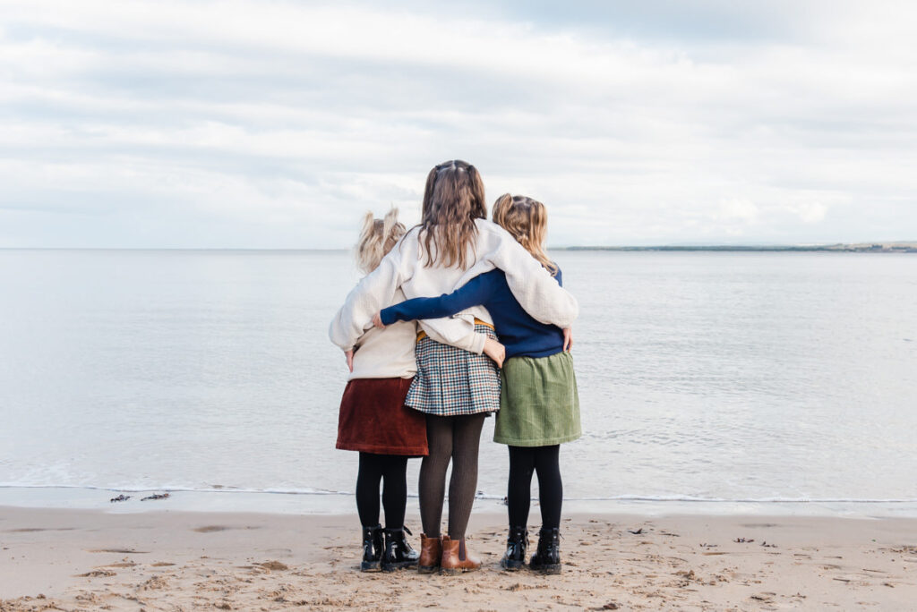 Three sisters stand arm in arm with their backs to the camera looking out to the sea during a Photo shoot on Roemarkie beach