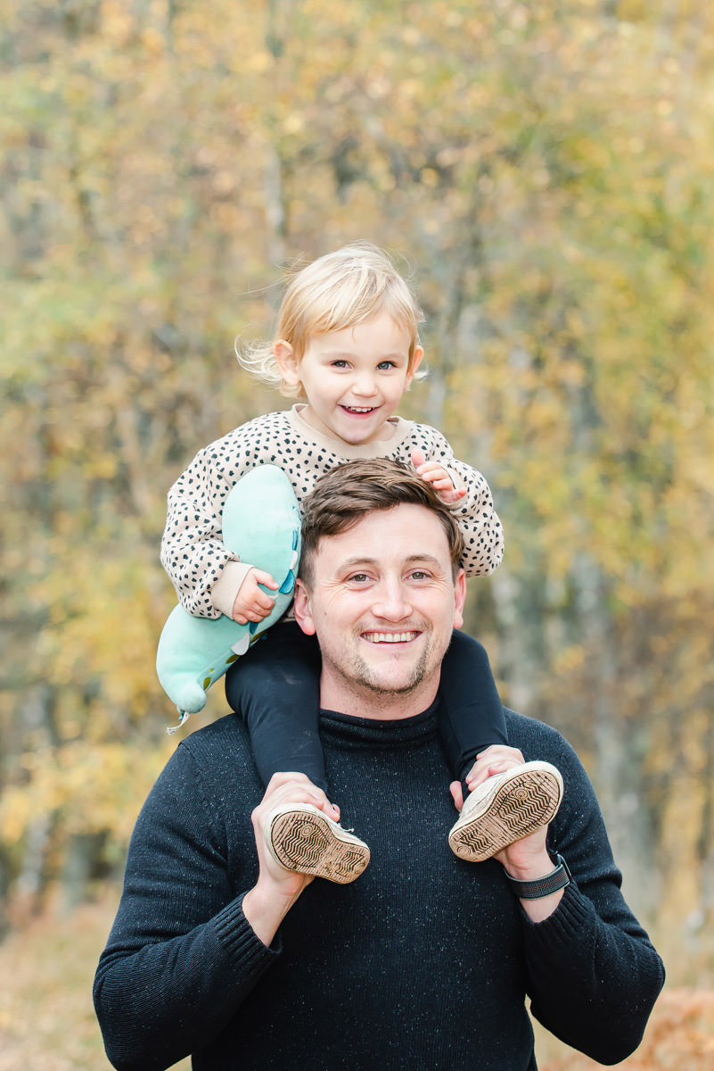 A young girl sits on her dads shoulders posing for a family photo shoot in a woodland setting in Aviemore