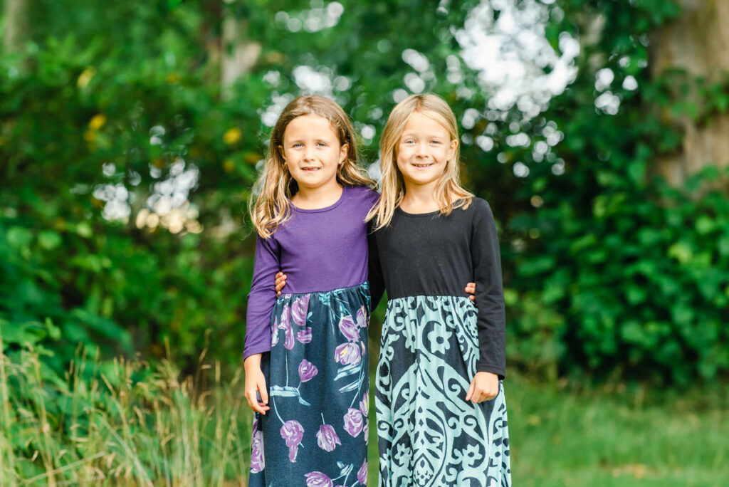 Two young sisters in long flowery dresses stand arm in arm as they pose for a Family Photo shoot in a woodland setting