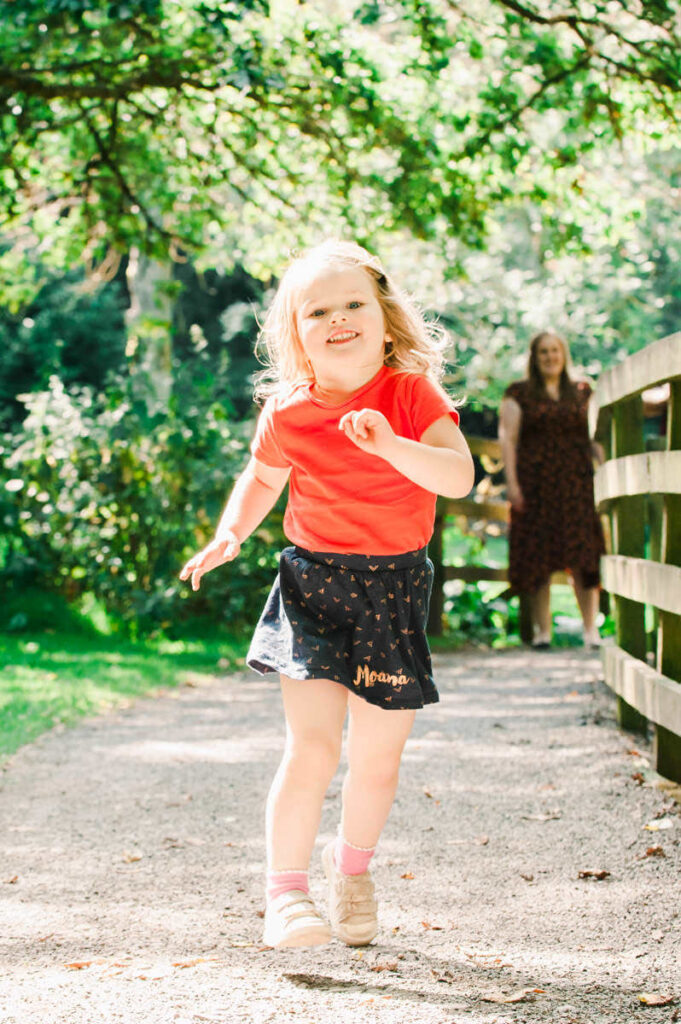 A young girl with blonde hair in a red top and a blue dress runs ahead as her mum watches on in a woodland setting