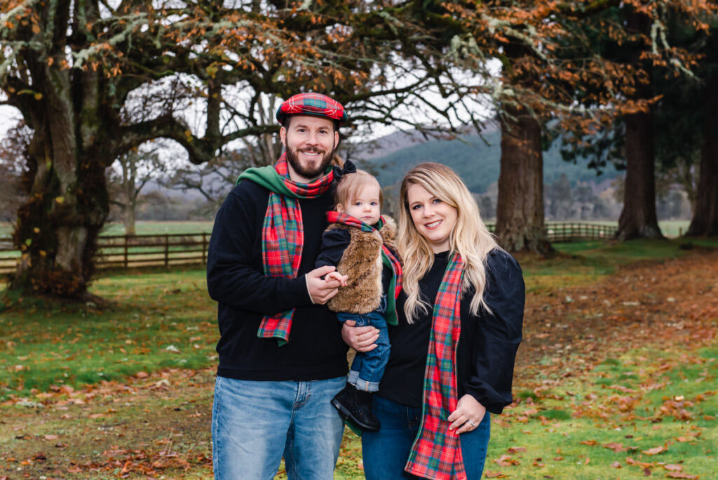 A family of three wearing tartan scarves and hats stand in a woodland setting in the grounds of Struy Castle