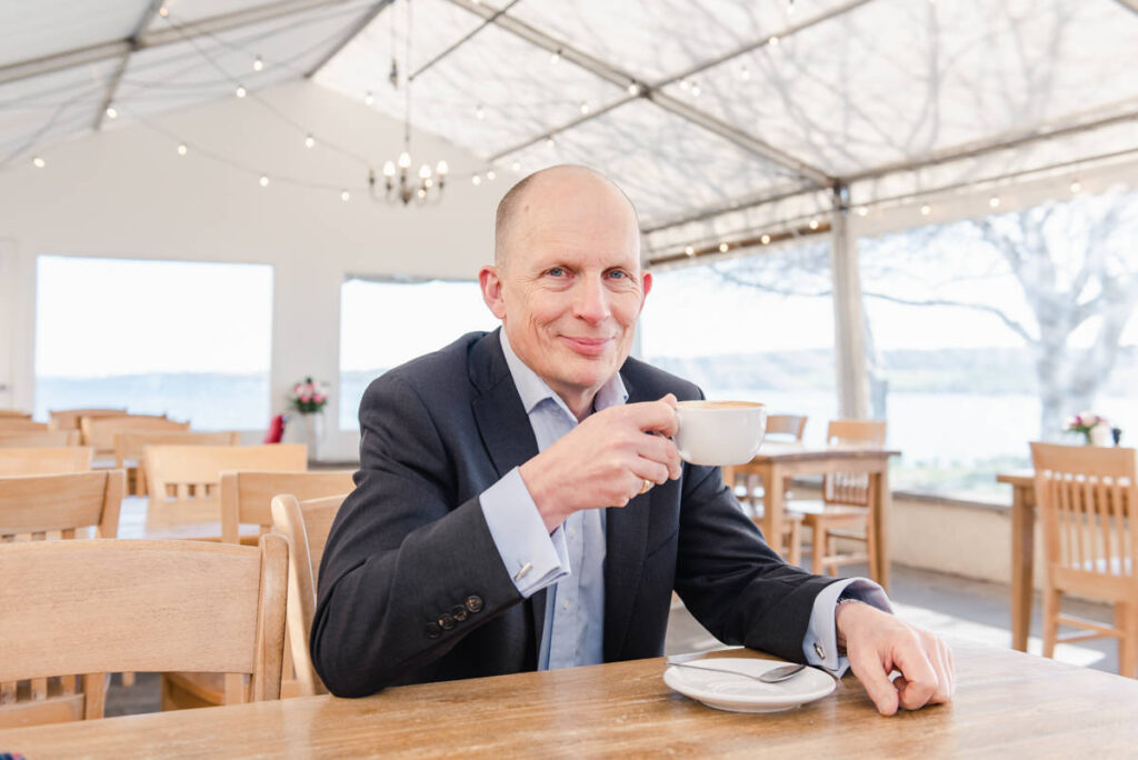 Black Isle dating profile head and shoulders portrait of a man in a jacket and shirt sitting at a table holding a coffee cup