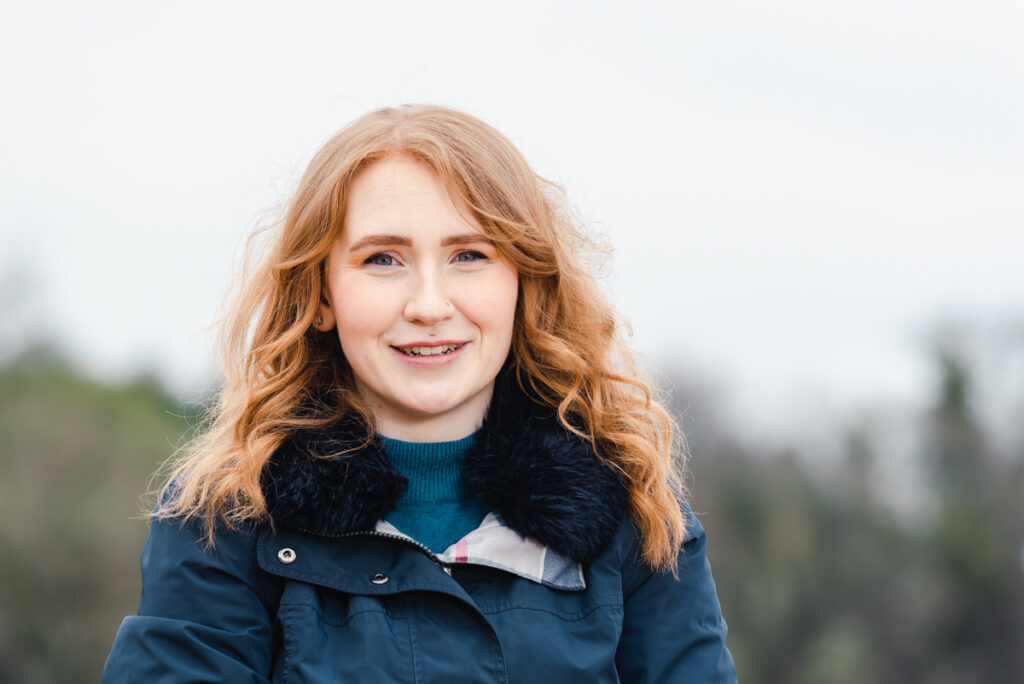 Outdoor headshot of a light-skinned woman with red hair and a navy jacket at Rosemarkie beach on the Black Isle