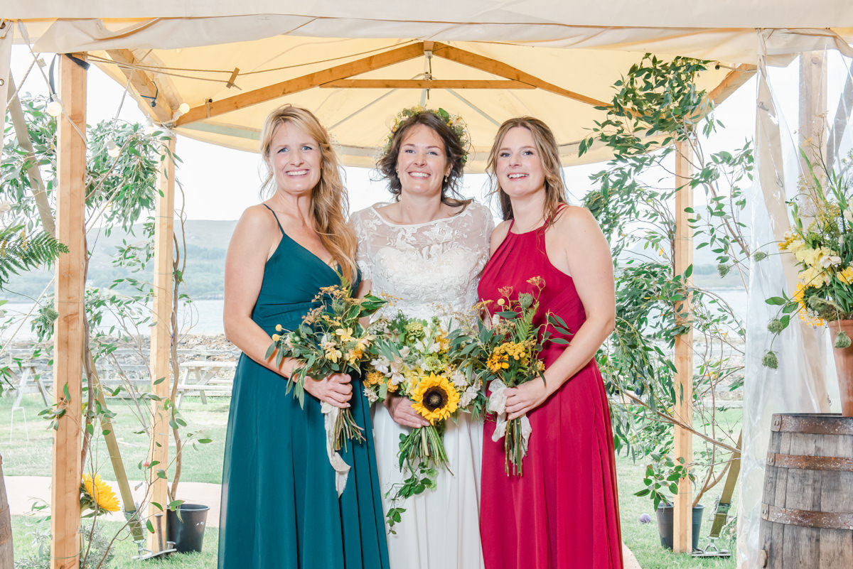 A bride wearing a floral headdress stands with her bridesmaids holding sunflower under a marquee in Ardmair near Ullapool