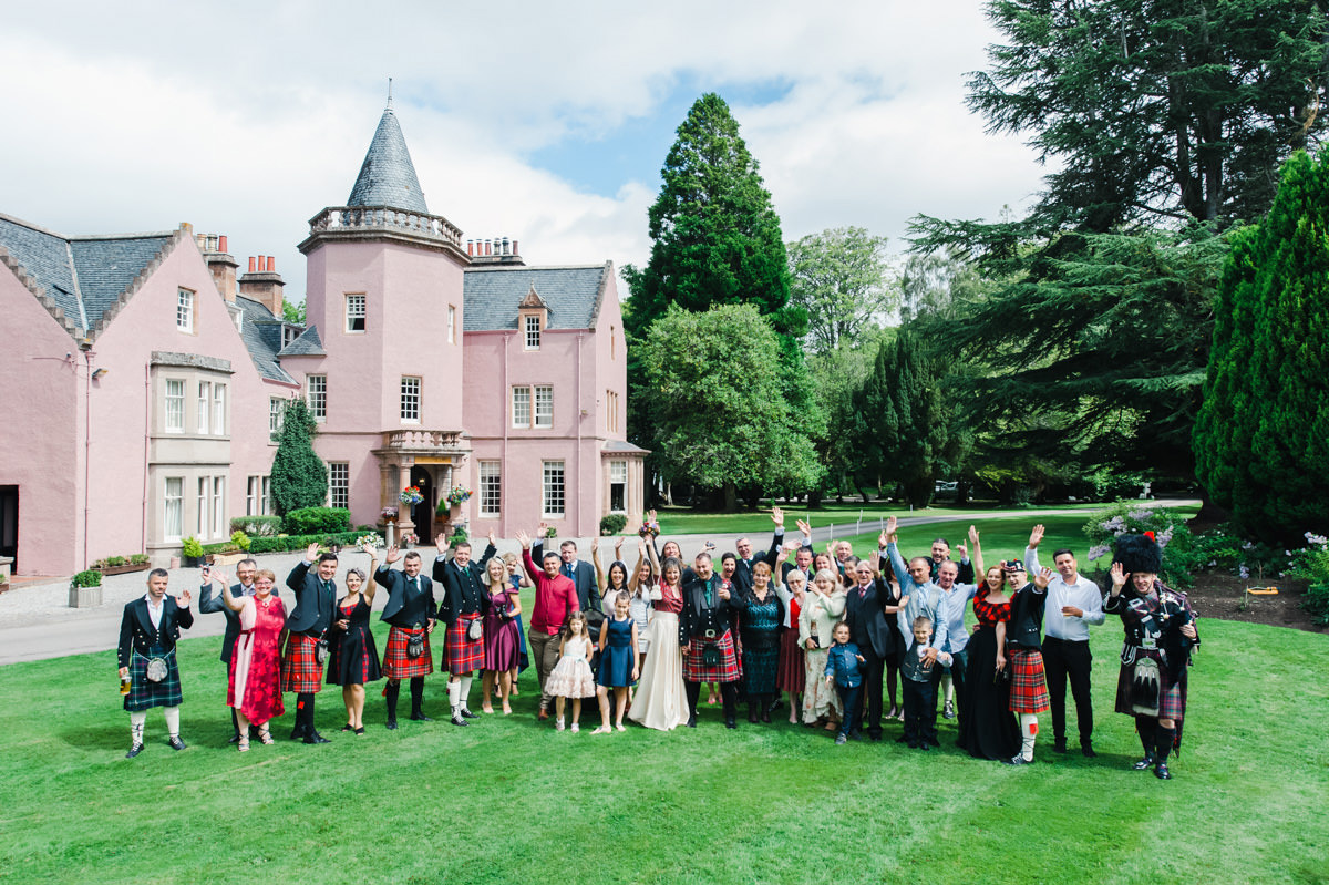 A large wedding party stand outside on the lawn of Bunchrew House waving at the camera