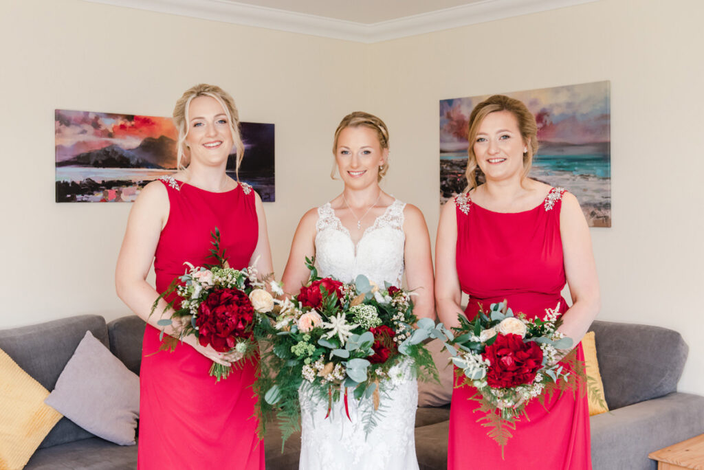 Two bridesmaids in pink stand either side of the bride holding large bouquets in a bright room near Loch Ness