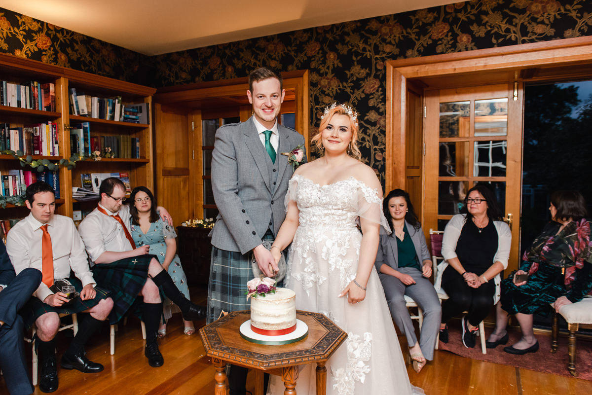 A bride and groom cut a single tiered wedding cake on a small wooden table while the guests watch in Embo House near Dornoch