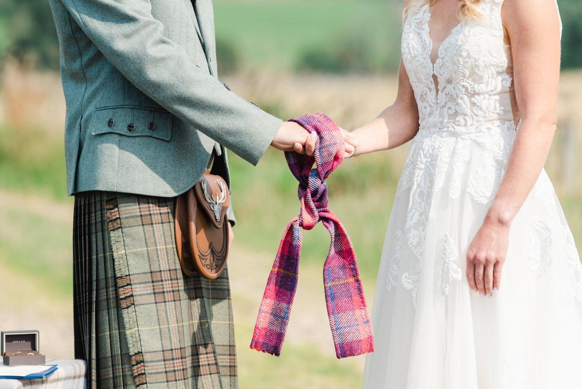 A groom and bride hold hands.A tartan cloth is also tied to their hands during a Handfasting Ceremony at an Ardross wedding