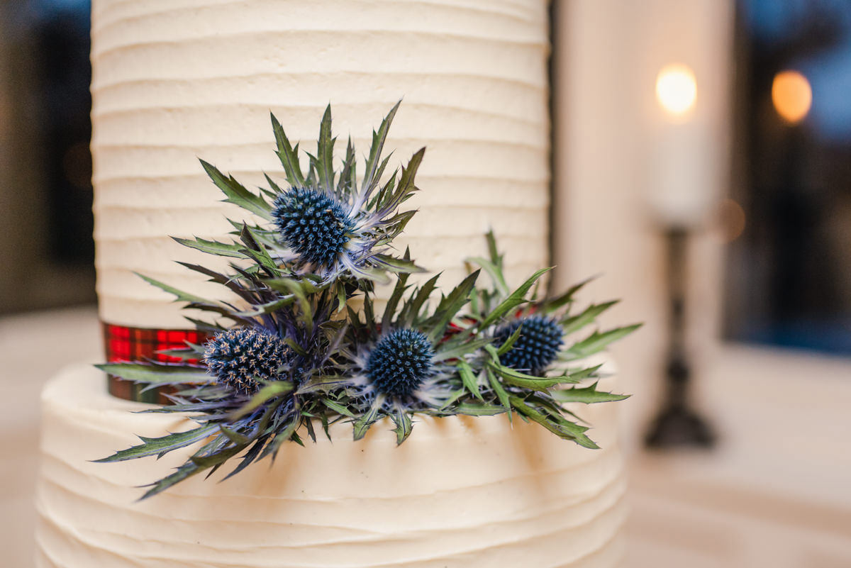 A close up of a thistle cake decoration with a tartan ribbon on a white wedding cake at the Invernairne Guest House
