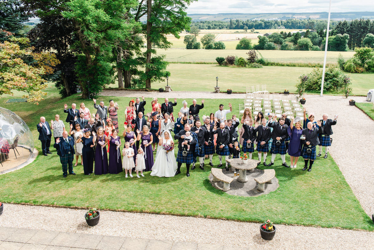 A wedding party stand on the lawn with trees behind them of Kincraig Castle Hotel looking upward and waving toward the camera