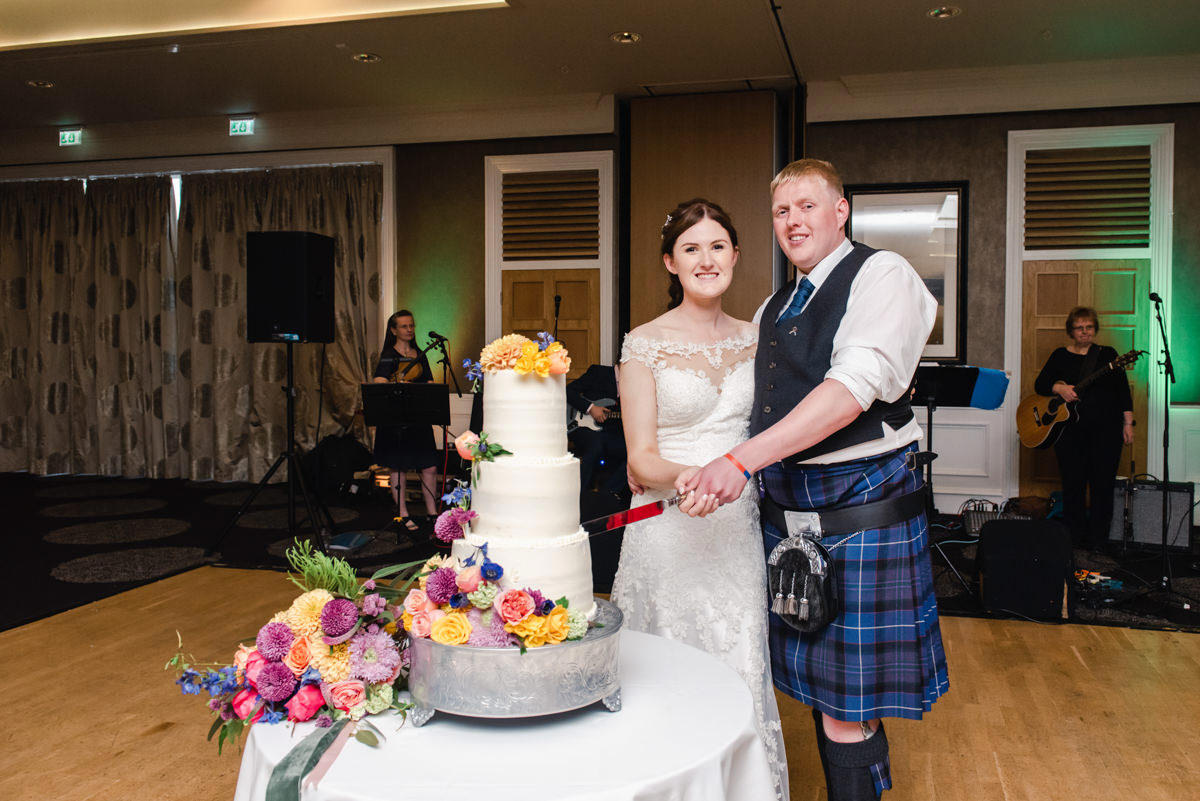 A bride and groom cut a white wedding cake on a white table with a floral display at its base in the Kingsmills Hotel
