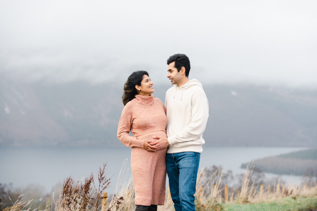A medium-skinned couple smiling at each other and holding the woman's pregnant belly on a couple's photoshoot at Loch Ness