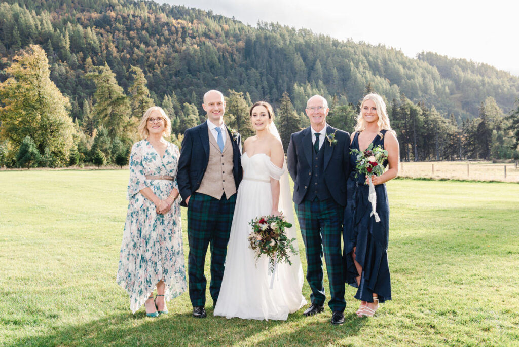 A wedding family group of five with the males dressed in tartan trousers stand in a field at Mar Lodge estate