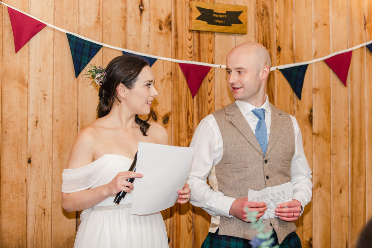 Bride and groom stand together with tartan and red bunting hanging behind them during their speech at a wedding in Mar Lodge