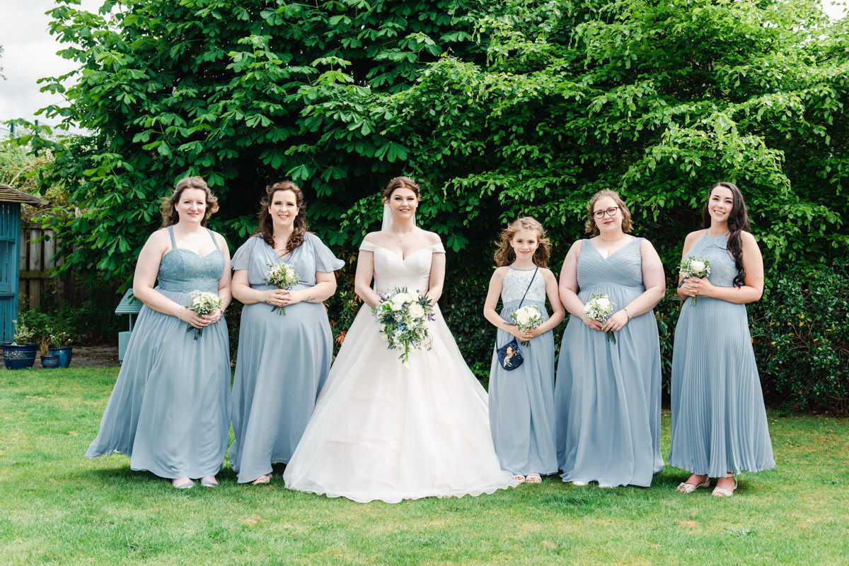 A bride in white stands in the centre of five bridesmaids dressed in lilac holding bouquets in front of a large conifer in Nairn