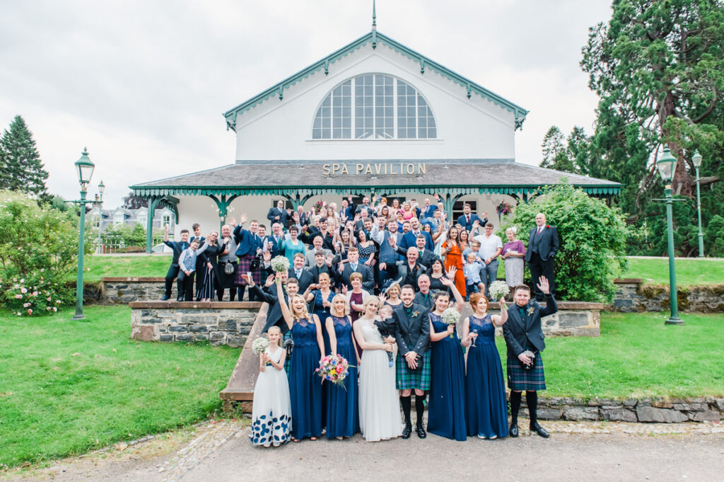 A large wedding party with bride and groom at the front, stands in front of Strathpeffer Pavilion waving at the camera