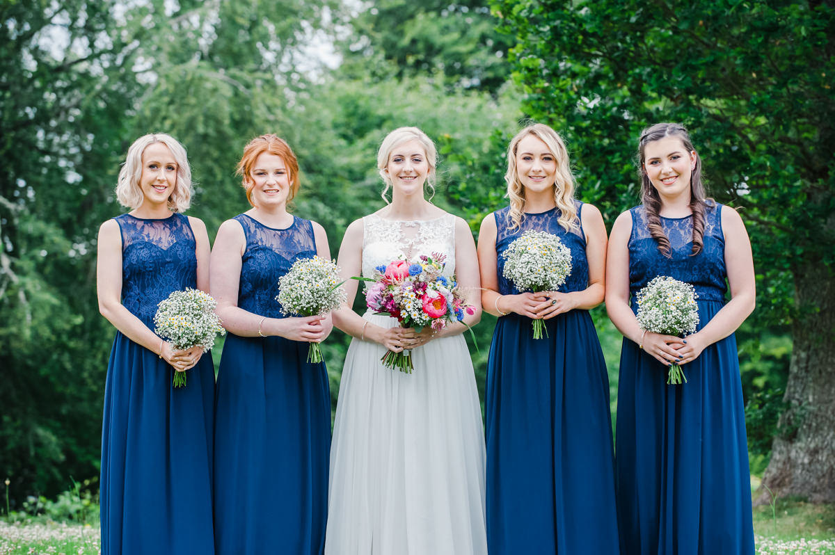 Bride in white standing with four bridesmaids in blue all holding bouquets in the gardens at Strathpeffer Pavilion
