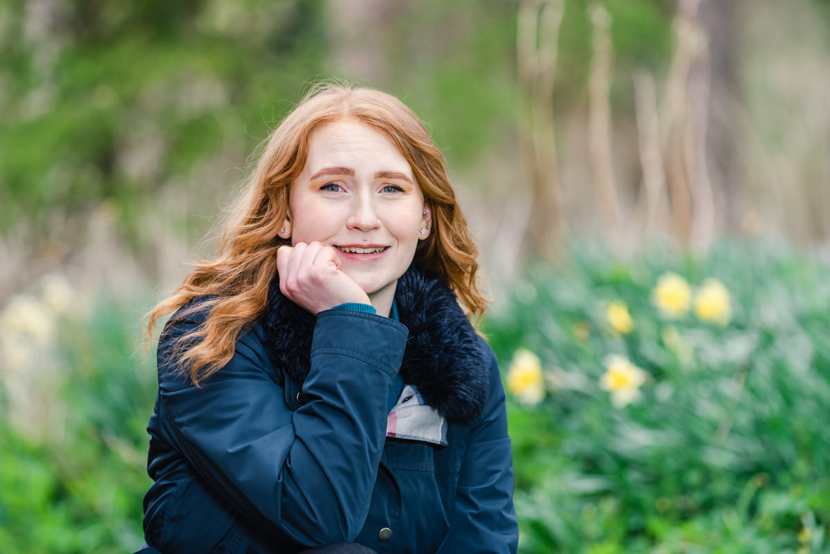 Outdoor portrait of a woman with ginger hair with her chin resting on her hand in front of daffodils in woods near Inverness