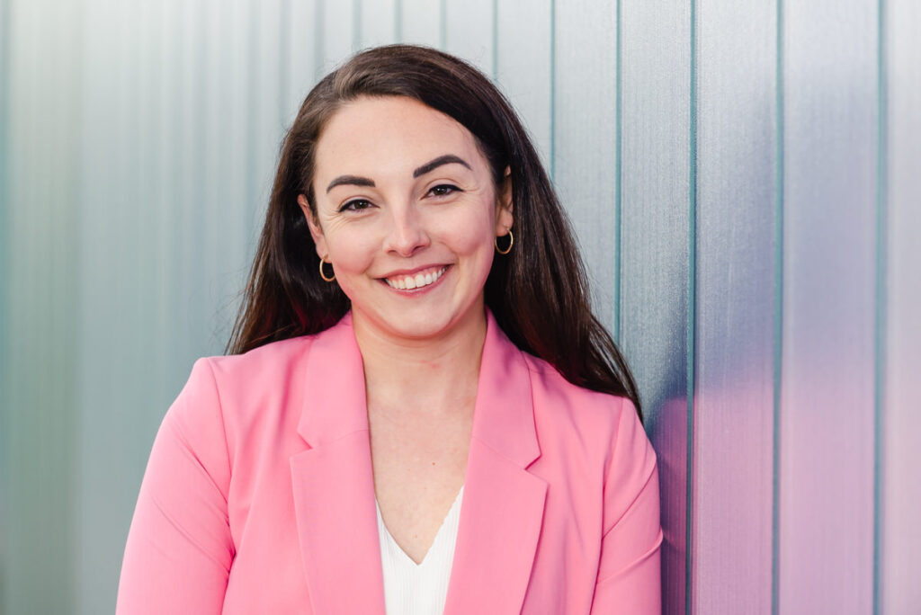 Professional headshot at Inverness Campus with a young woman with long brown hair and a pink blazer leaning on a purple wall