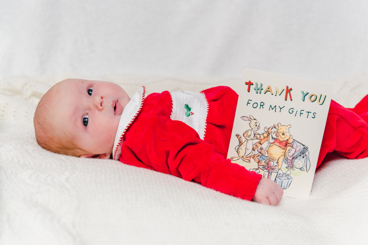 A red headed baby wearing a red jumpsuit lying on a white bed with a white background looking at the camera