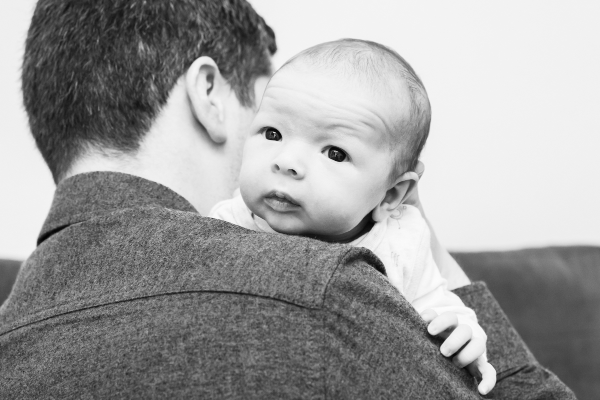 A black and white image of a newborn looking over its fathers shoulder while being cuddled