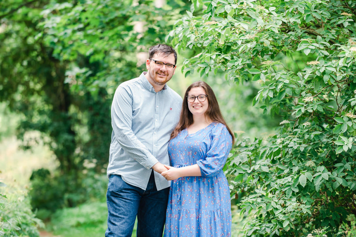 In a deciduous woodland setting a casually dressed young couple wearing glasses hold hands and smile toward the camera