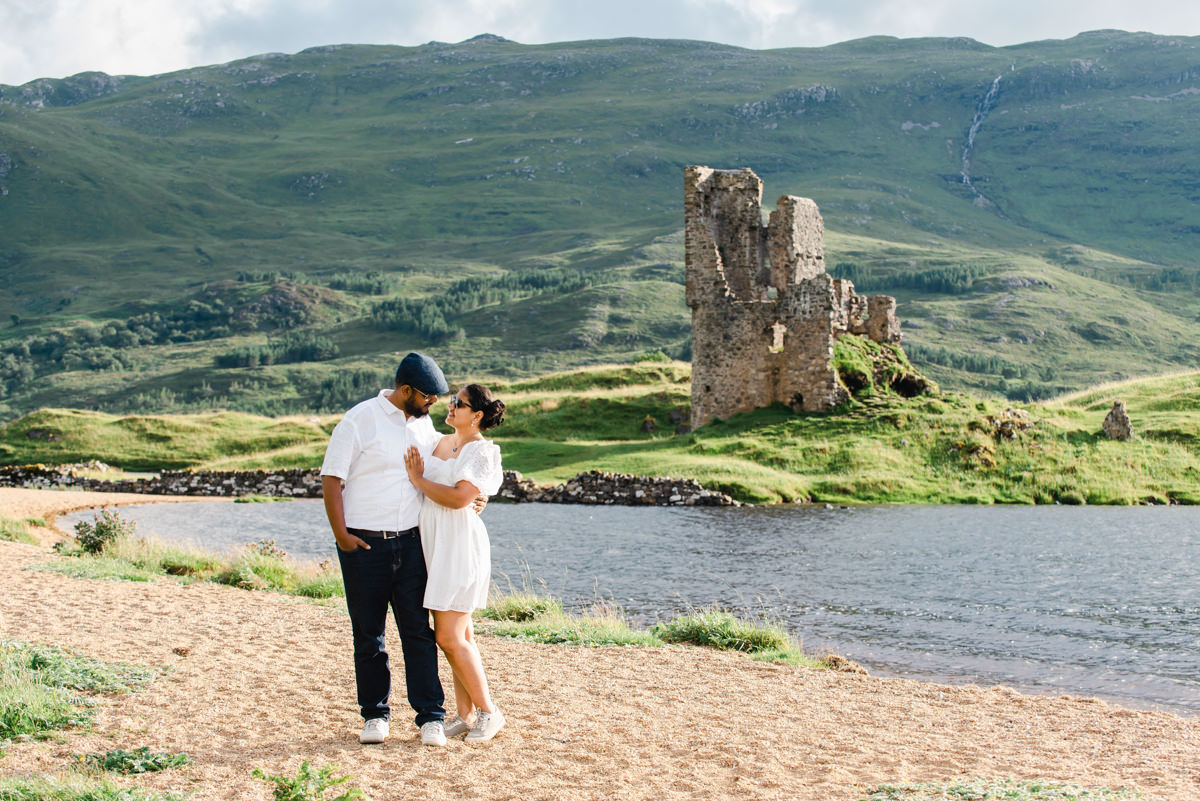 A young dark skinned couple in white share a tender moment while standing on a beach next to a ruined Scottish castle