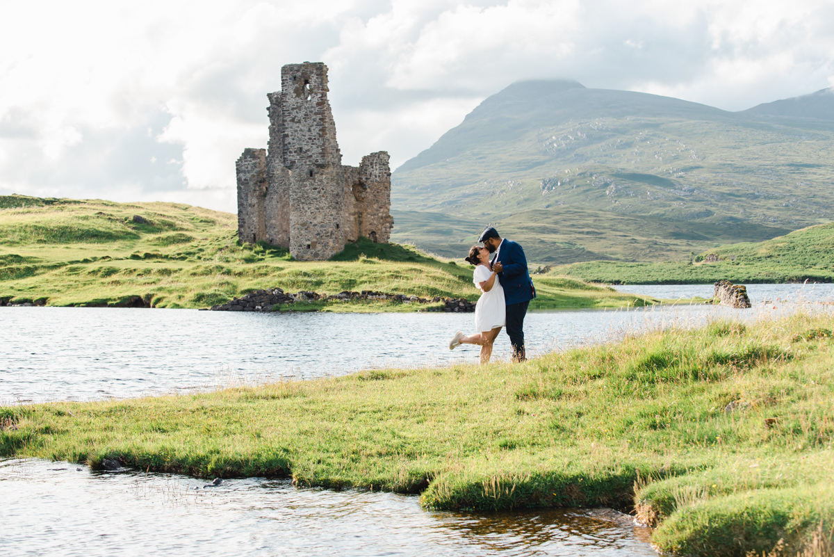 At the side of a loch with a ruined Scottish castle behind them a young dark skinned couple share an affectionate kiss