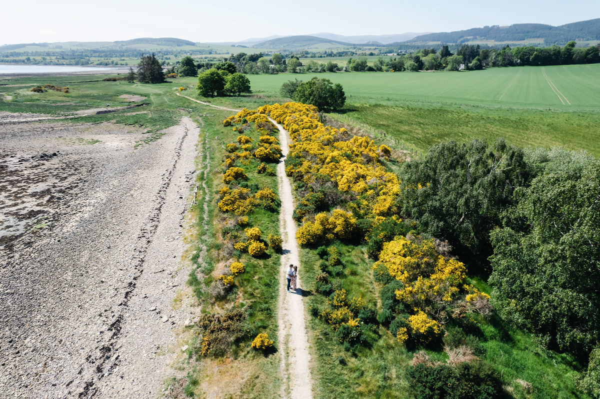 A birds-eye view of a couple embracing and kissing on a track with yellow gorse either side by a stony beach