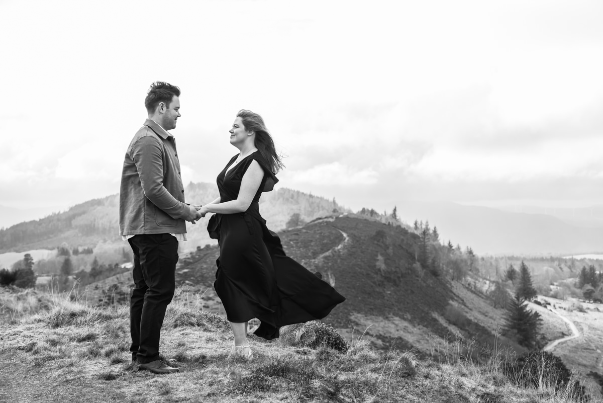 A black and white image of a couple facing each other and holding hands on a hilltop as the wind blows the females dark dress