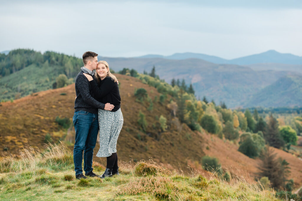 On a hilltop overlooking a scene of conifer woodlands and heather a couple embrace as the male kisses the head of the female