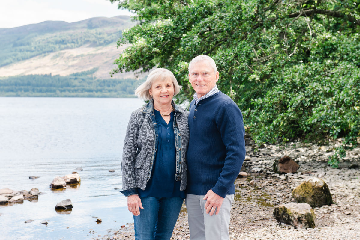 A mature couple dressed in blue stand side by side on the stony beach of a loch with over hanging alder tress to the side
