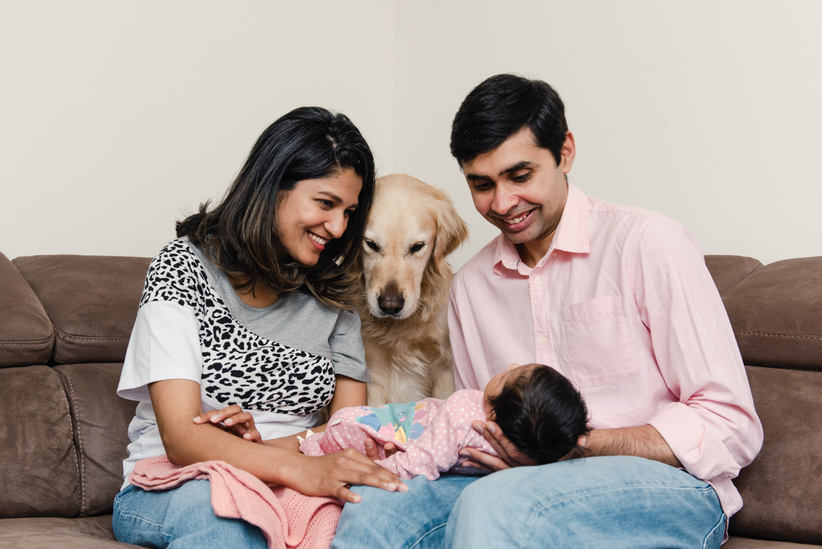 A young couple and their dog sit together on a beige sofa while lovingly holding their newborn baby