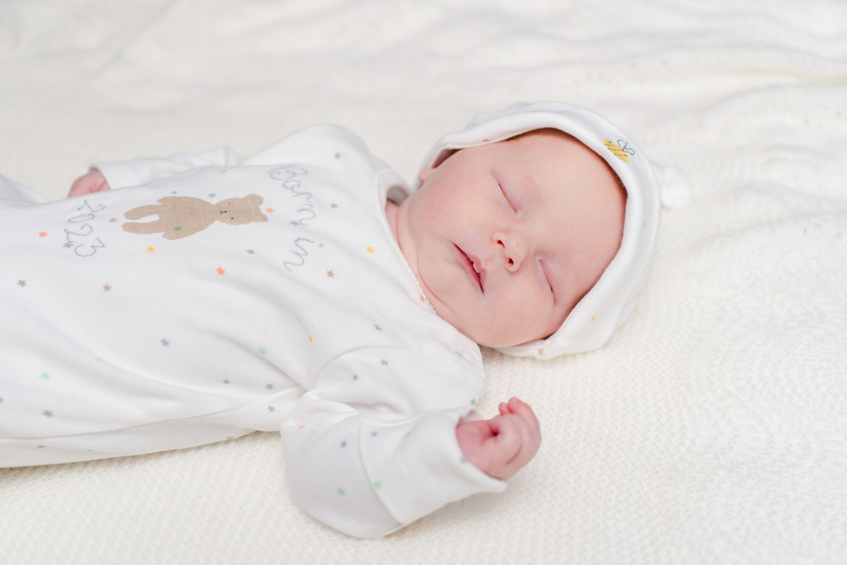 A sleeping newborn baby in a white onesie and white hat lays on its back on white bedclothes