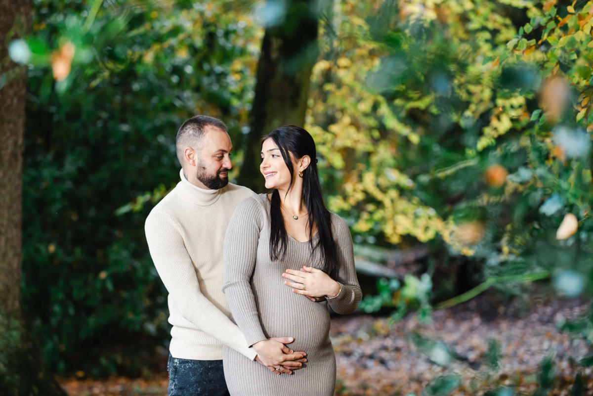 In a green woodland setting a couple look at each other while both holding the baby bump