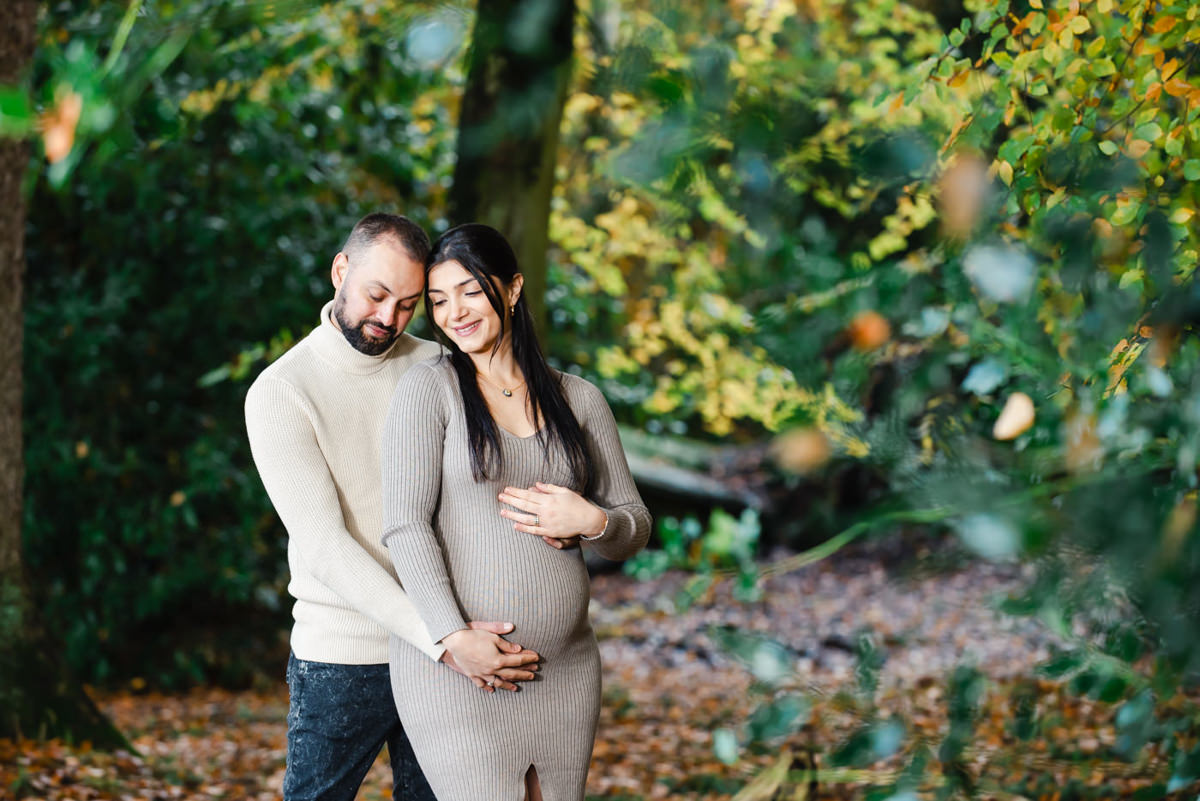 In a woodland setting a young couple with eyes closed hold each others hands over the mothers baby bump