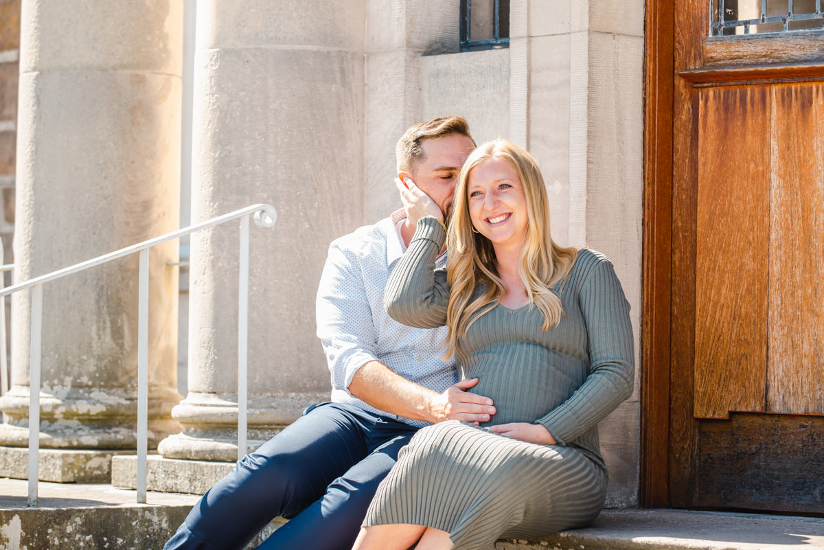 Seated on the steps of a sandstone building a blonde pregnant woman lovingly cups her partners face as he holds the baby bump