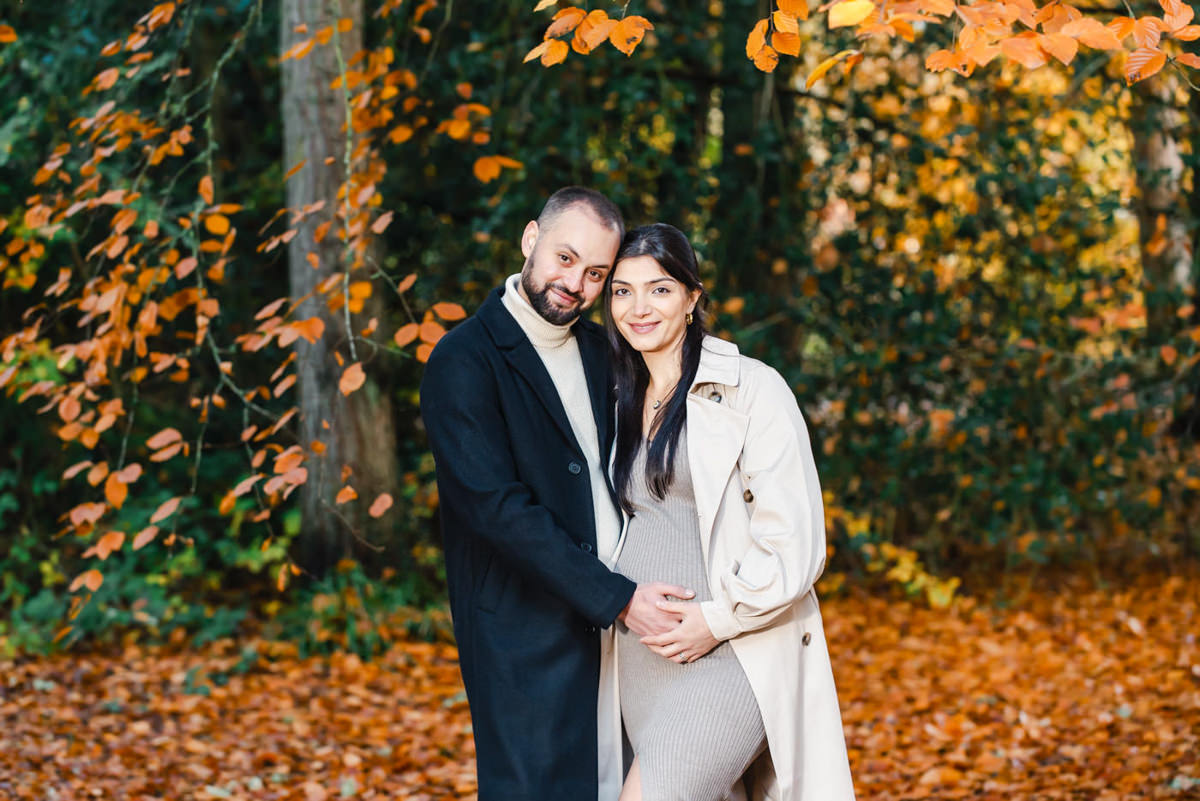 In an autumnal beech tree setting a young couple together hold the mothers baby bump and smile affectionately at the camera