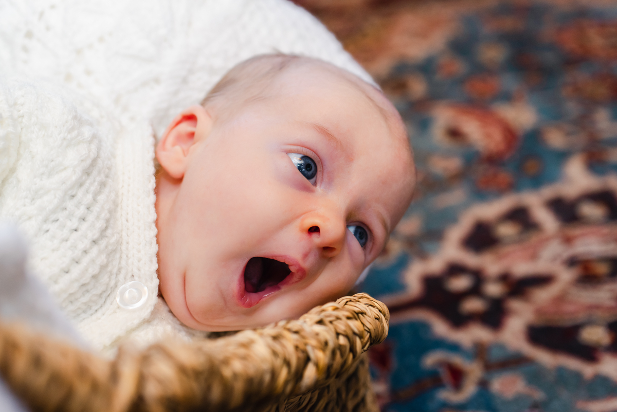 A close up of a yawning baby in a Moses basket dressed in a white woolen cardigan