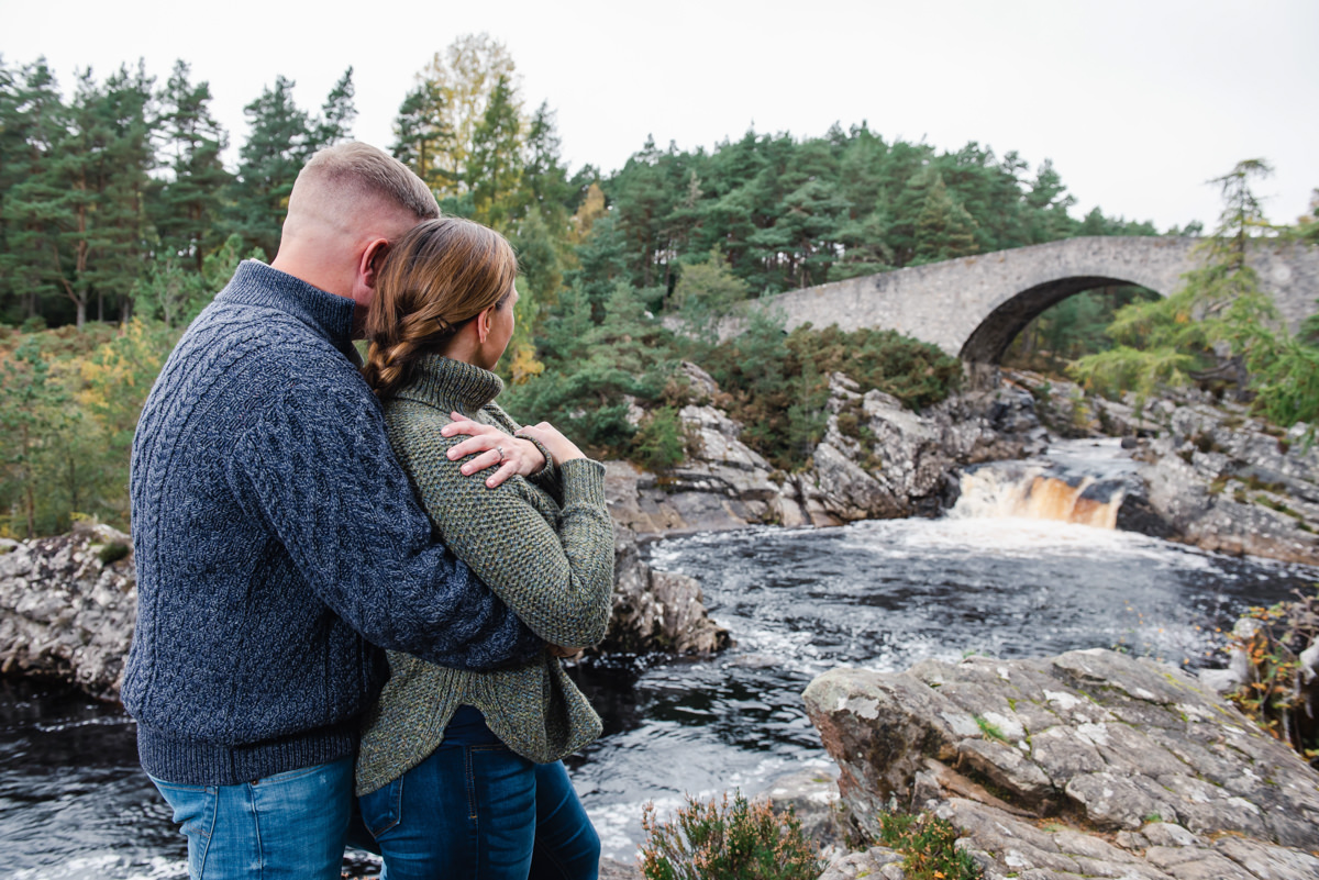 A couple in woolen jumpers embrace while looking toward an old stone bridge and gushing dark river surrounded by conifer trees