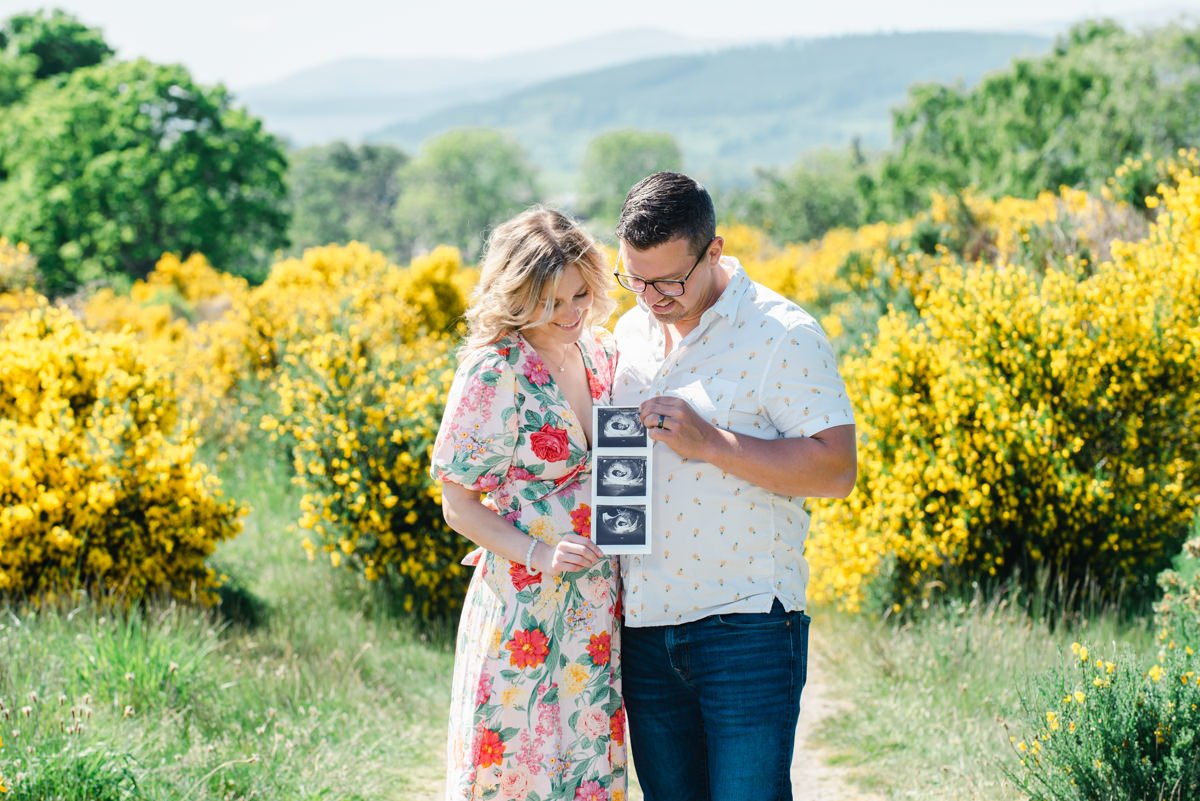 A couple stand on a summers day in front of bright yellow gorse bushes while holding scans of there new born baby to be